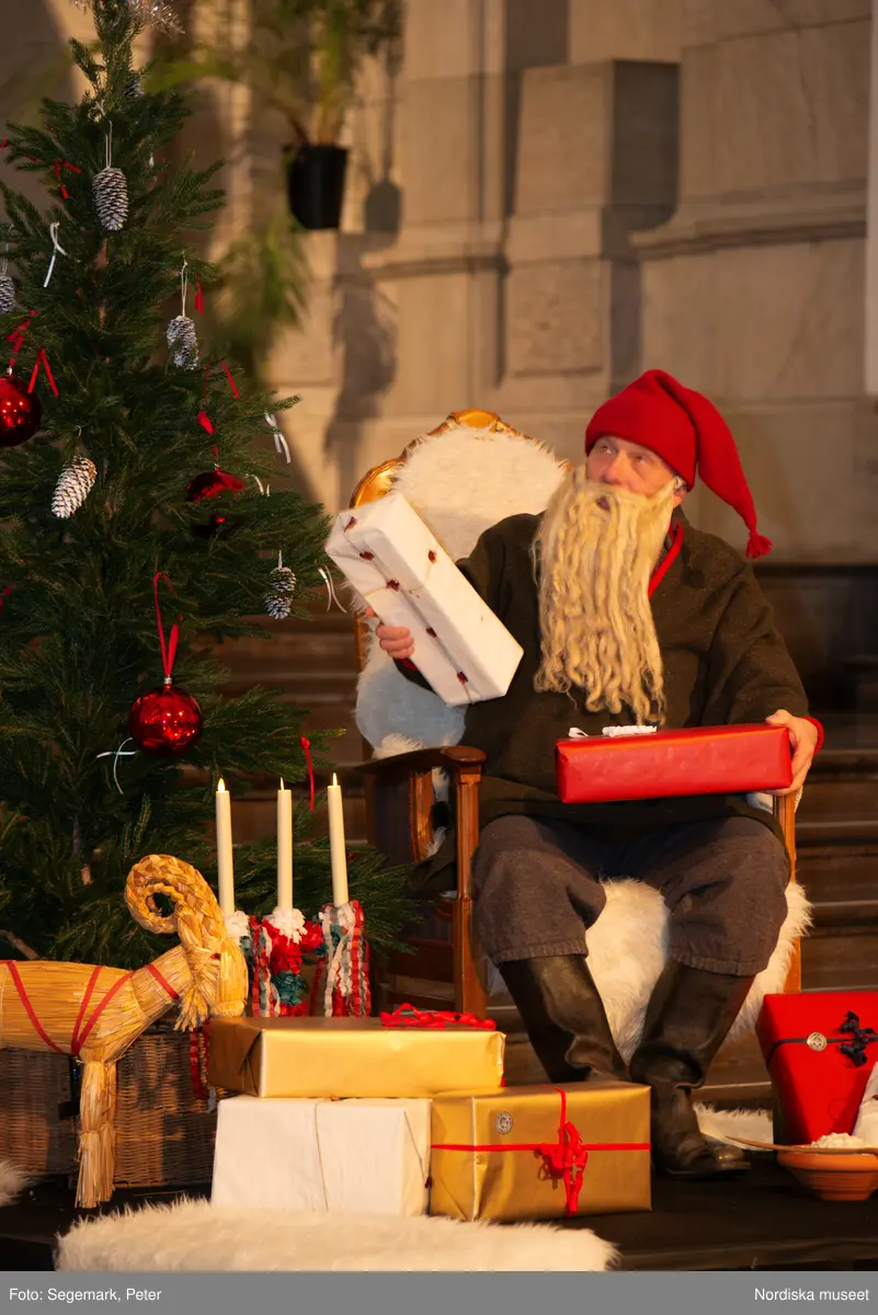 En tomte är på besök i Nordiska museet. Han sitter vid den klädda julgranen och läser på julklapparna. Nedanför granen inslagna paket och en julbock.