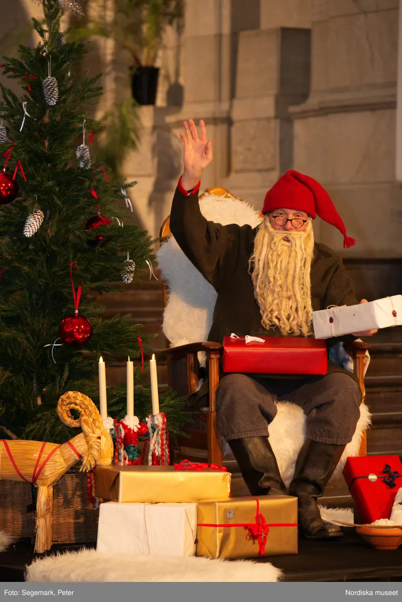En tomte är på besök i Nordiska museet. Han sitter vid den klädda julgranen och läser på julklapparna. Nedanför granen inslagna paket och en julbock.