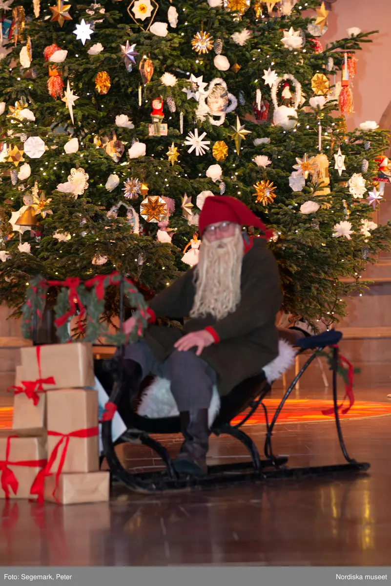 En tomte är på besök i Nordiska museet. Han sitter vid den klädda julgranen och läser på julklapparna. Nedanför granen inslagna paket och en julbock.