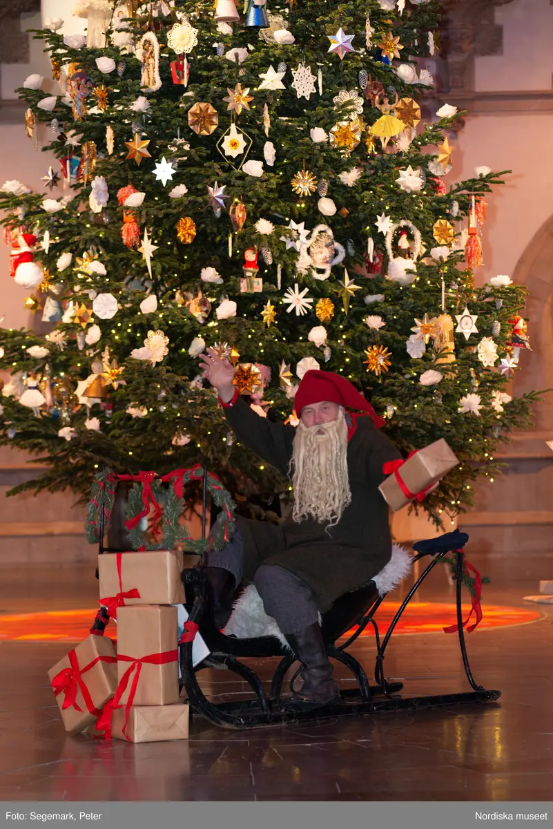 En tomte är på besök i Nordiska museet. Han sitter vid den klädda julgranen och läser på julklapparna. Nedanför granen inslagna paket och en julbock.