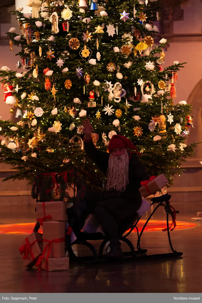 En tomte är på besök i Nordiska museet. Han sitter vid den klädda julgranen och läser på julklapparna. Nedanför granen inslagna paket och en julbock.