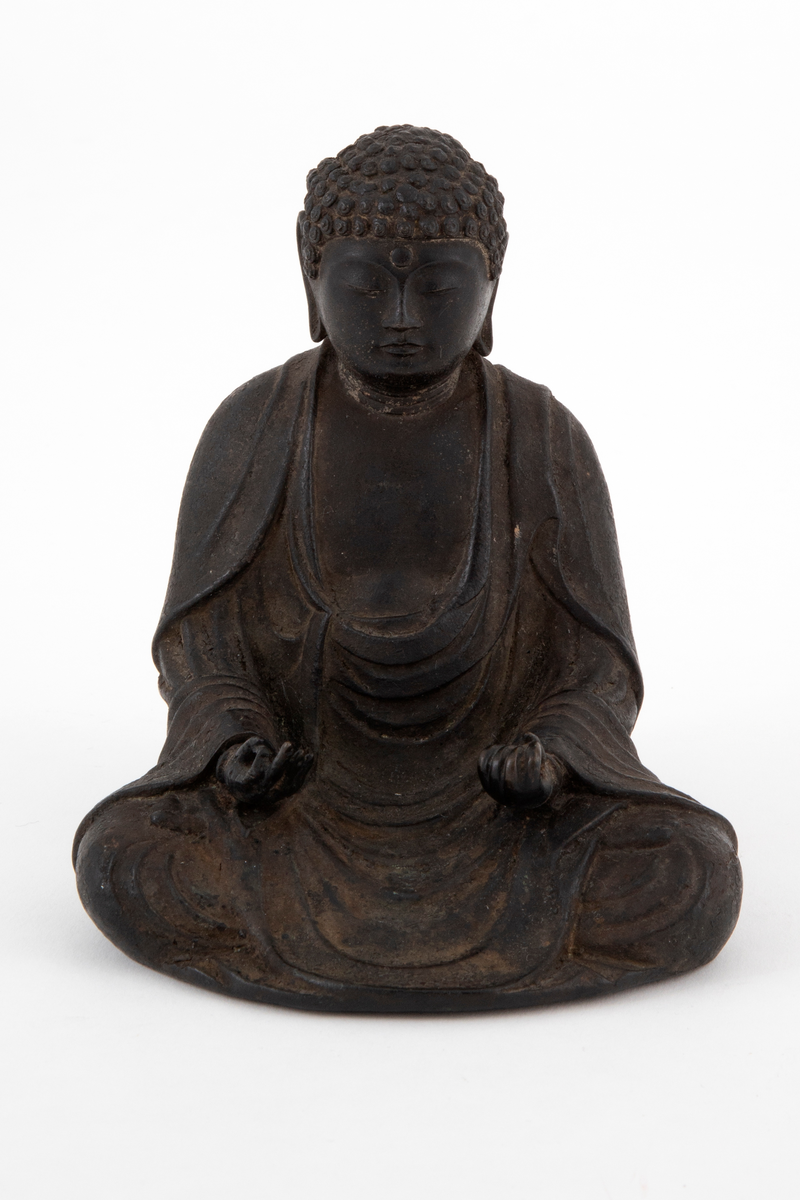 Støpt bronsefigur forestillende Buddha sittende i en meditativ stilling med bena i kors, såkalt lotusstilling. Figuren er hul innvendig. På ryggen en bøyle.