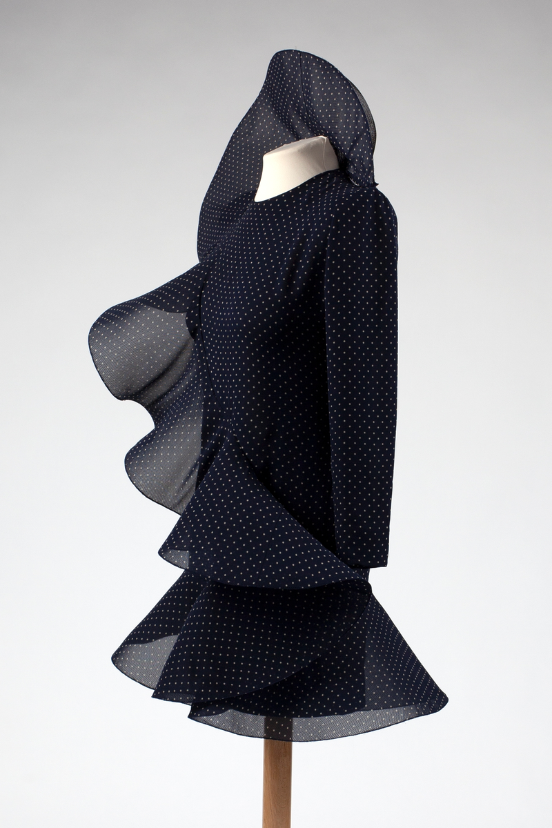 Figurnær, nesten lårkort cocktailkjole av midnattsblå, delvis transparent chiffon med små, hvite prikker, såkalte "polkadotter". Brede, rette skuldre, meget lange, trange ermer, lukning med glidelås midt bak. En påsydd, bred krage, er kneppet på venstre skuldersøm, og reiser seg som en halvsirkel bak hodet, før den bølger ned som en volang diagonalt over kjolens front, for å ende i i en struttende kappe nederst på kjolen. Både krage, volang og kappe er avstivet med et stivt nettstoff av syntetmateriale.

I Christie's auksjonskatalog er kjolen avbildet med nedbrettet krage, men i motemagasinet L'Officiel, er den avbildet i 1991. med stående krage.