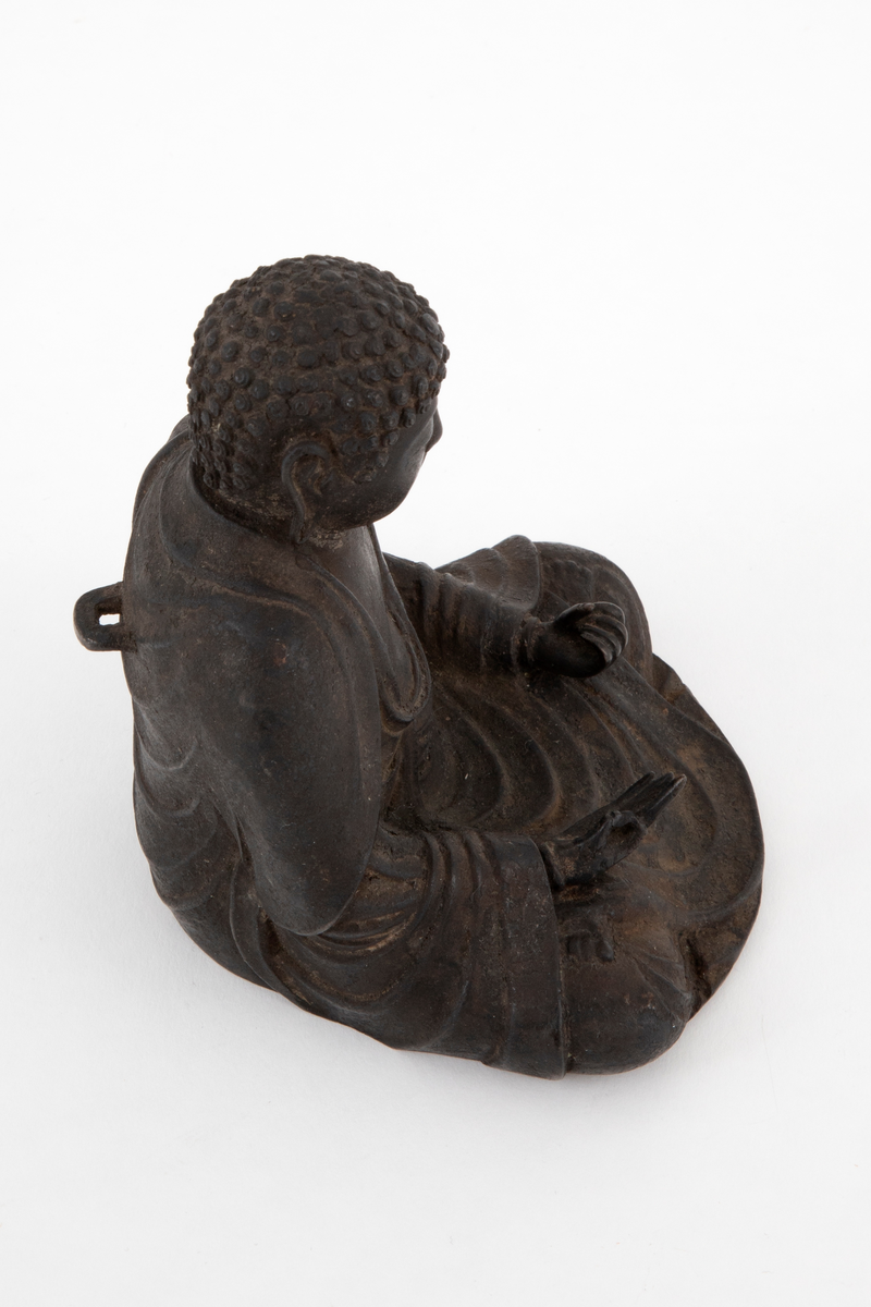 Støpt bronsefigur forestillende Buddha sittende i en meditativ stilling med bena i kors, såkalt lotusstilling. Figuren er hul innvendig. På ryggen en bøyle.