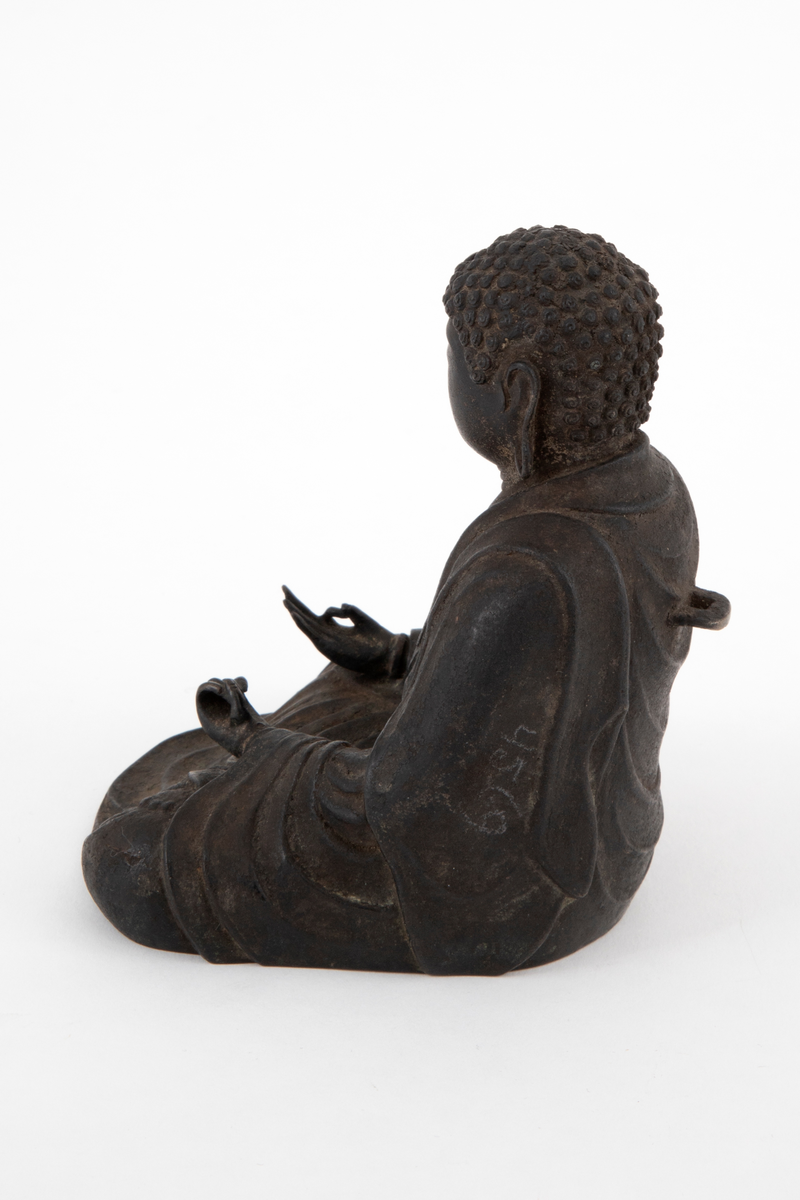 Støpt bronsefigur forestillende Buddha sittende i en meditativ stilling med bena i kors, såkalt lotusstilling. Figuren er hul innvendig. På ryggen en bøyle.