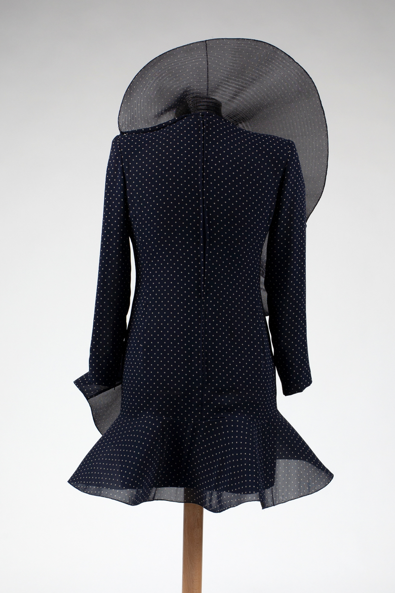 Figurnær, nesten lårkort cocktailkjole av midnattsblå, delvis transparent chiffon med små, hvite prikker, såkalte "polkadotter". Brede, rette skuldre, meget lange, trange ermer, lukning med glidelås midt bak. En påsydd, bred krage, er kneppet på venstre skuldersøm, og reiser seg som en halvsirkel bak hodet, før den bølger ned som en volang diagonalt over kjolens front, for å ende i i en struttende kappe nederst på kjolen. Både krage, volang og kappe er avstivet med et stivt nettstoff av syntetmateriale.

I Christie's auksjonskatalog er kjolen avbildet med nedbrettet krage, men i motemagasinet L'Officiel, er den avbildet i 1991. med stående krage.
