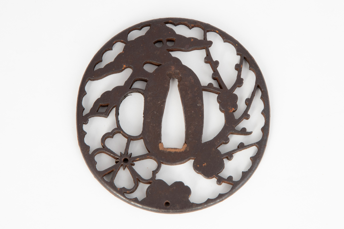 Sirkelrund parerplate, tsuba, i jern. I gjennombrutt motiv vises grener, blomster og furukroner i positiv silhuett. Jernet er patinert sort.