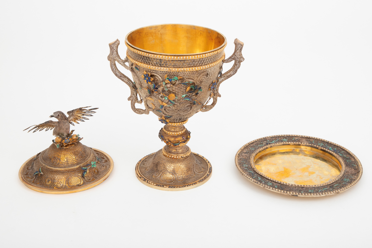 Pokal med lokk og fat utført i forgylt metall. Rund, hvelvet fotplate, balusterformet stett, begerformet kupa samt hvelvet, profilert lokk. Pokal og lokk er overtrukket av et tett nett i filigran. Kupa har to hanker formet som naturalistiske grener i filigran. Herfra brer kvister med blomster, blad og fugler seg over kupa. Blomstene og flere av bladene er i blå, gul, lilla og grønn emalje. Lignende dekor på lokket og stettens utbukting. Perlerader på stetten og øvre del av kupa. På lokkets topp står en fugl med utspilte vinger i filigran. Pokalen står på et rundt fat med flat bunn og flat kant. Kanten er dekorert med to perlerader mellom hvilke det er et filigransnett med blomster og blader i grønn og blå emalje.