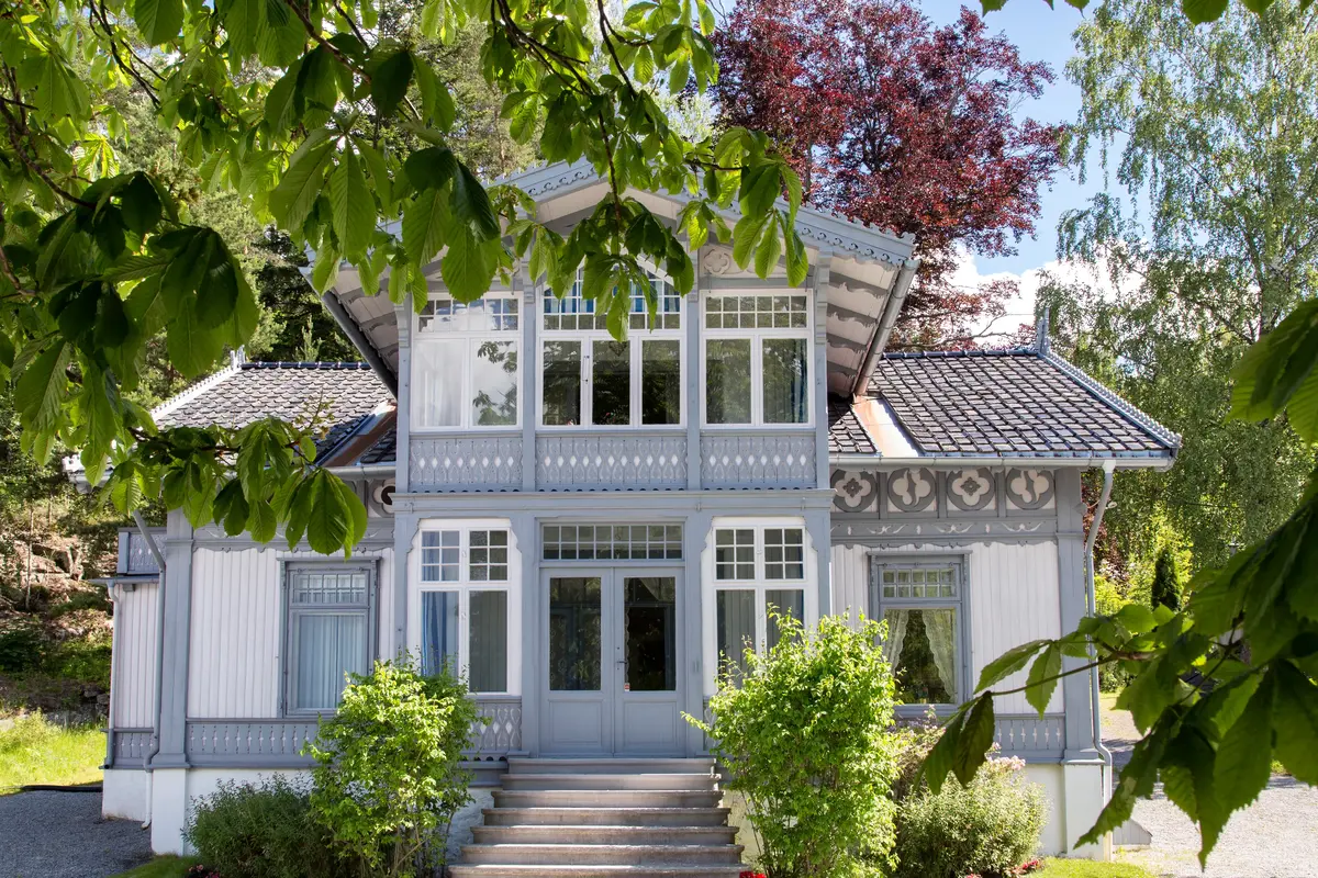 Eldre trehus i hvitt og grått med fine utskjæringer, med trær og grønne planter rundt.