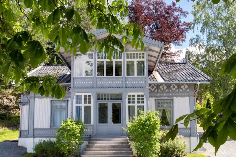 Eldre trehus i hvitt og grått med fine utskjæringer, med trær og grønne planter rundt.