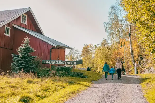Låve  til venstre med skilt foran, tre mennesker som går på en sti på høyre side.