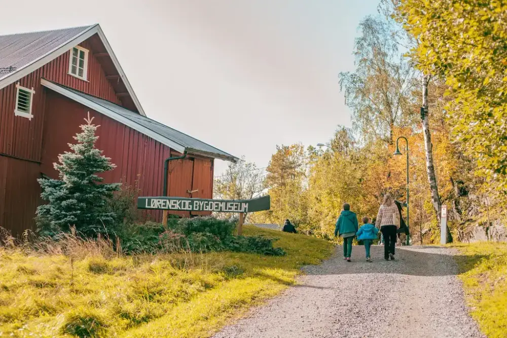 Låve  til venstre med skilt foran, tre mennesker som går på en sti på høyre side.