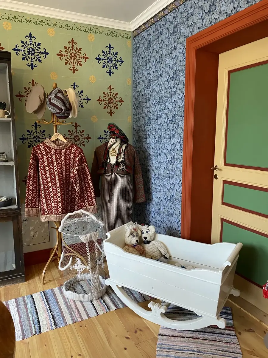 Hörna i ett rum. Blå tapet på den ena väggen och den andra väggen är grön med mönster i blå, röd och gult. På den blå väggen finns en dörr som går i rött, grönt och en vitaktig färg. På golvet står en utstyrsel på en mannekäng, en hatthängare med hattar och en stickad rödvit tröja och en vit vagga med två nallebjörnar. Två små trasmattor på golvet. 