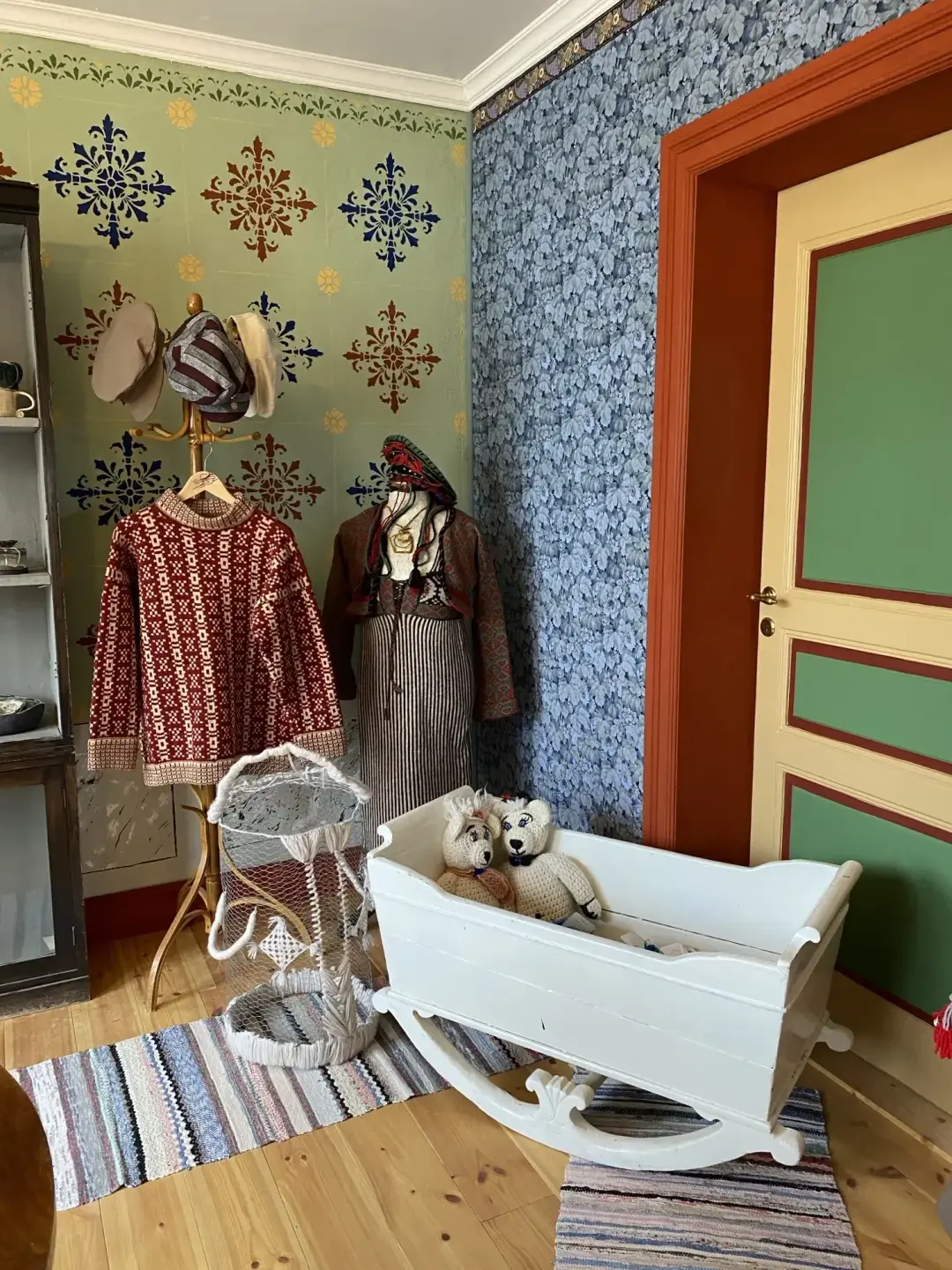 Hörna i ett rum. Blå tapet på den ena väggen och den andra väggen är grön med mönster i blå, röd och gult. På den blå väggen finns en dörr som går i rött, grönt och en vitaktig färg. På golvet står en utstyrsel på en mannekäng, en hatthängare med hattar och en stickad rödvit tröja och en vit vagga med två nallebjörnar. Två små trasmattor på golvet. 