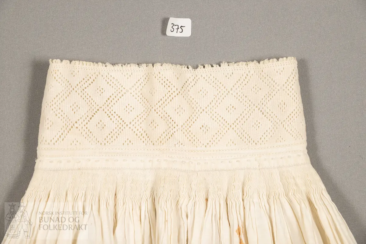 Halvskjorte, sauma av tynn, fin linlerret i framstykker og krage. Bomullslerret i nakkestykket. Brodert stolpe framme med hardangersaum. Nakkestykket striperynka og sydd til krage. Kragen brodert med uttrekksaum.