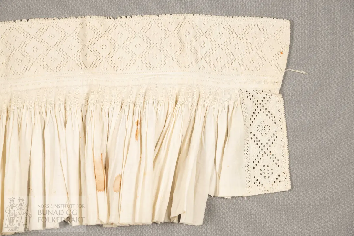Halvskjorte, sauma av tynn, fin linlerret i framstykker og krage. Bomullslerret i nakkestykket. Brodert stolpe framme med hardangersaum. Nakkestykket striperynka og sydd til krage. Kragen brodert med uttrekksaum.