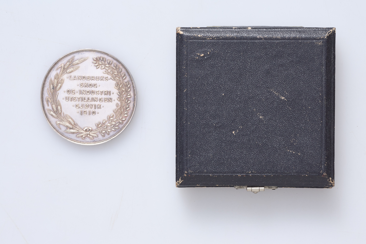 Medalje (a) med etui (b). Medaljen er fra landbruks-, skog- og industriutstillingen på Gjøvik i 1910. Etuiet er fra J. Frisenberg, Lillehammer.