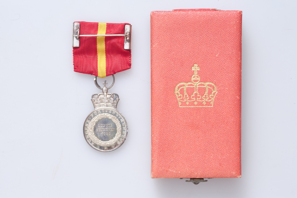 Kongens fortjenestmedalje i sølv (a) med etui (b). Medaljen er festet i et rødt og gult bånd. Tildelt Ludvig Skjerven i 1960. Etuiet dekorert med krone på lokket.