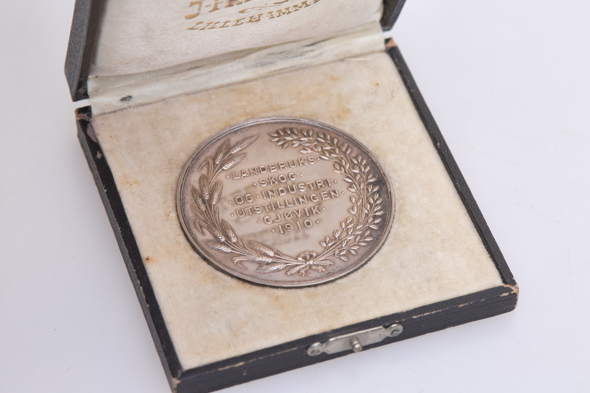 Medalje (a) med etui (b). Medaljen er fra landbruks-, skog- og industriutstillingen på Gjøvik i 1910. Etuiet er fra J. Frisenberg, Lillehammer.