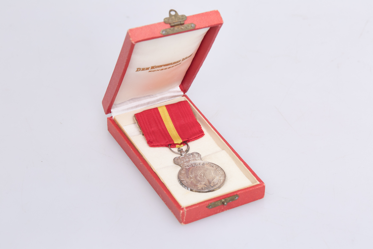 Kongens fortjenestmedalje i sølv (a) med etui (b). Medaljen er festet i et rødt og gult bånd. Tildelt Ludvig Skjerven i 1960. Etuiet dekorert med krone på lokket.