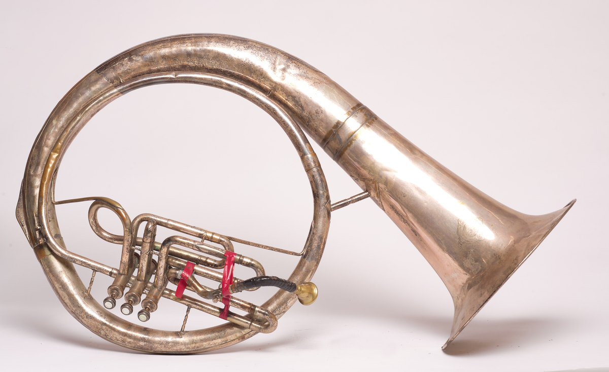 Ess-tuba produsert av Brødrene Schuster i Markneukirchen, Tyskland. Blåse - ventilinstrument. Sjalstykke med to ringer, når og tre ventiler. Bæres. Bygget i ring, rundt hals på skuldrer.