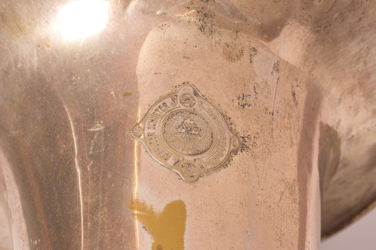 Ess-tuba produsert av Brødrene Schuster i Markneukirchen, Tyskland. Blåse - ventilinstrument. Sjalstykke med to ringer, når og tre ventiler. Bæres. Bygget i ring, rundt hals på skuldrer.