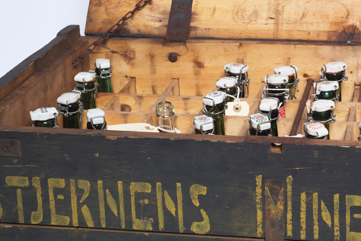 Seltersflaske med patentkork. Korken er merket med "Rena". Flasken er oppbevart sammen med flere andre flasker i en trekiste med lokk. På trekisten står det "Stjernens Mineralvande" på siden.