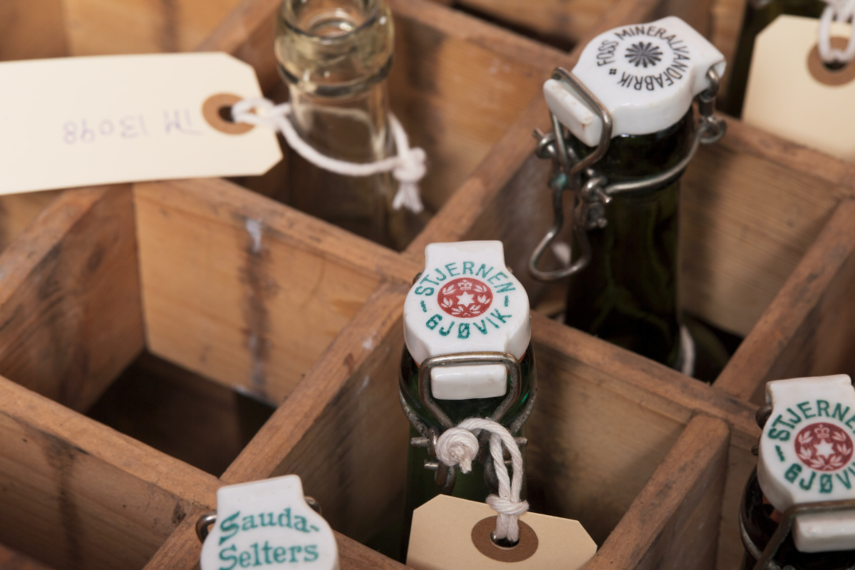 Seltersflaske med patentkork. Korken er merket med "Rena". Flasken er oppbevart sammen med flere andre flasker i en trekiste med lokk. På trekisten står det "Stjernens Mineralvande" på siden.