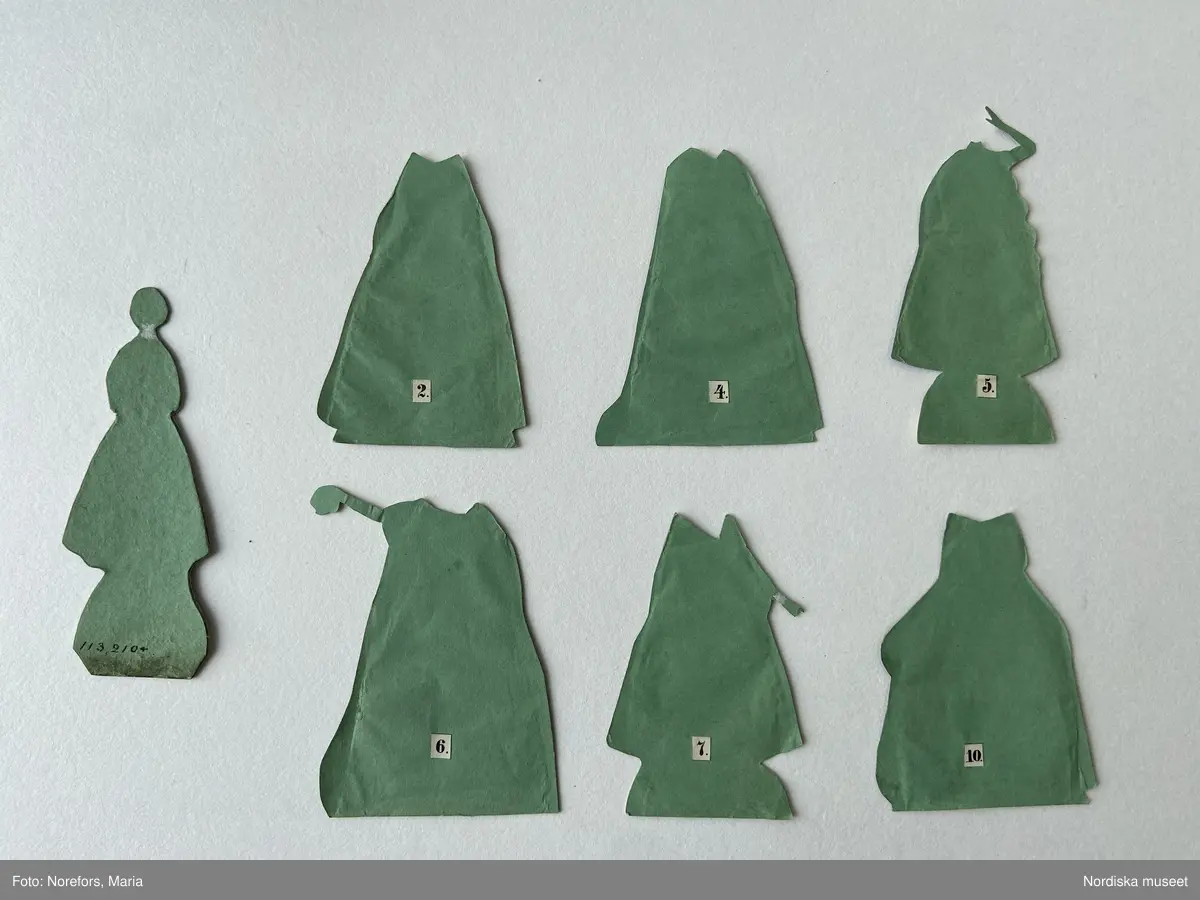 Inventering Sesam 1996-1999:
H 17, B 10,8 cm (kartongen)
Pappersdocka av kartong fodrad med grönt papper, tryck och handkolorerad, föreställande den svenska sångerskan Jenny Lind (1820-1887), som slog igenom stort på 1840-tal. Dockan ligger i sin originalkartong tillsammans med en fotställning av kork och tio kolorerade dräkter.
Dockan (H 10 cm) har gul-brunt uppsatt hår och är klädd i en underkjol och ett snörliv i vitt, vita strumpor och svarta skor snörda upp på vaderna. Hon håller armarna i kors över bröstet. Originalkartongen består av en underdel och ett separat lock, materialet är kartong, grön undertill, rosa på sidorna och ovanpå en guldpappersram med en kolorerad bild av Jenny Lind i röd klänning med spetskrage och hennes namn skrivet som överskrift. På var sida om bilden står skrivet på tyska respektive engelska tio operor och huvudroller som Jenny Lind sjungit och som kläderna i kartongen hör till.
Operor, roller och tillhörande dräkt:
1. Robert the Devil, rollen som Alice, klädd i en brun dräkt, vitt band runt midjan med ett radband och en vandringsstav.
2. The Huguenots, rollen som Valentine, klädd i en röd klänning med vida ärmar och spetskrage.
3. The somnambule, rollen som Amine, klädd i folkdräktslik klädsel med vit blus, blått snörliv och gul kjol med röda ränder.
4. Don Juan, rollen som Donna Anna, klädd i vit klänning och svart mantel.
5. The daughter of the regiment (Regementets dotter), rollen som marketenterskan Mary, klädd i en vit uniformsjacka och en blå kjol, bärande på en öltunna.
6. Norma, rollen som Norma, klädd i en vit klänning och en blå mantel.
7-8. Vielka, rollen som Vielka, klädd i en duvblå och gul klänning med vackra broderier och vida ärmar, samt en dress med rött liv med gulddekoration och en mörkblå kjol.
9. The Freisch¿tz, rollen som Agathe, klädd i en blå överdel, ett rosa band runt midjan och en gul kjol.
10. konsertklänning, blekrosa klänning med spetskrage och ytterligare en vit kjol under. 
Se "Leka för livet", Nordiska museets och Skansens årsbok, Fataburen 1992, Uddevalla 1991. Avsnittet "Lyckan att klä av och på en docka", av Ingrid Roos Björklund, s.109ff.
Helena Carlsson 1996