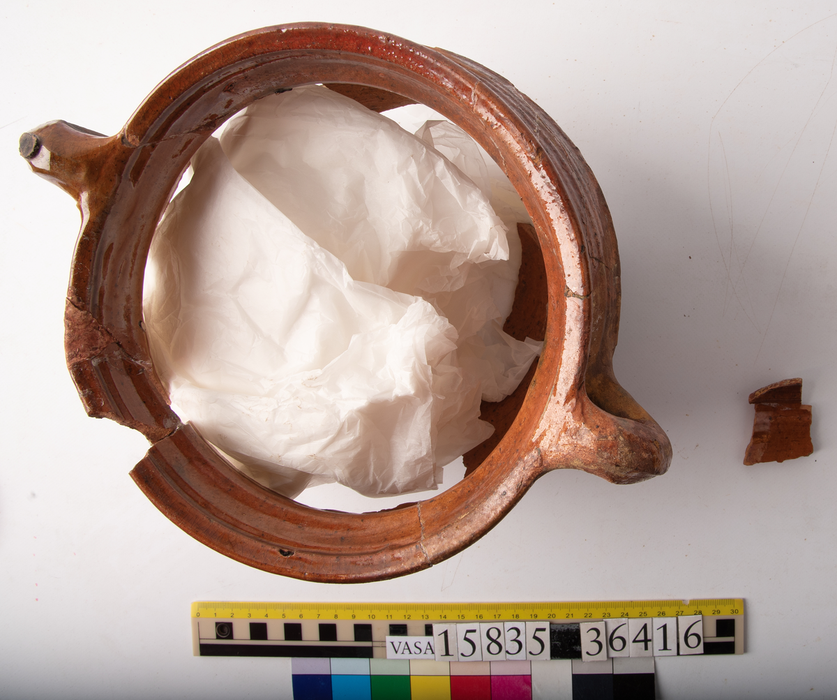 Kruka med två trindhänklar, ej komplett.
Krukan är fragmentarisk och sammansatt av ett flertal delar och bitar. Hela botten och delar av buken borta. Mynningsranden och bröstet nästan komplett. Mynningsvidd 190 mm. Förtjockad och profilerad mynningsrand med två trindhänklar som är ca 10 mm högre än mynningskanten.
På bröstet drejfåror. På skudervinkeln en ås och på bukkanten en ås.
Krukan är in- och utvändigt brunt blyglaserad, på buken mycket dåligt glaserad där den verkar sotig och sönderbränd.