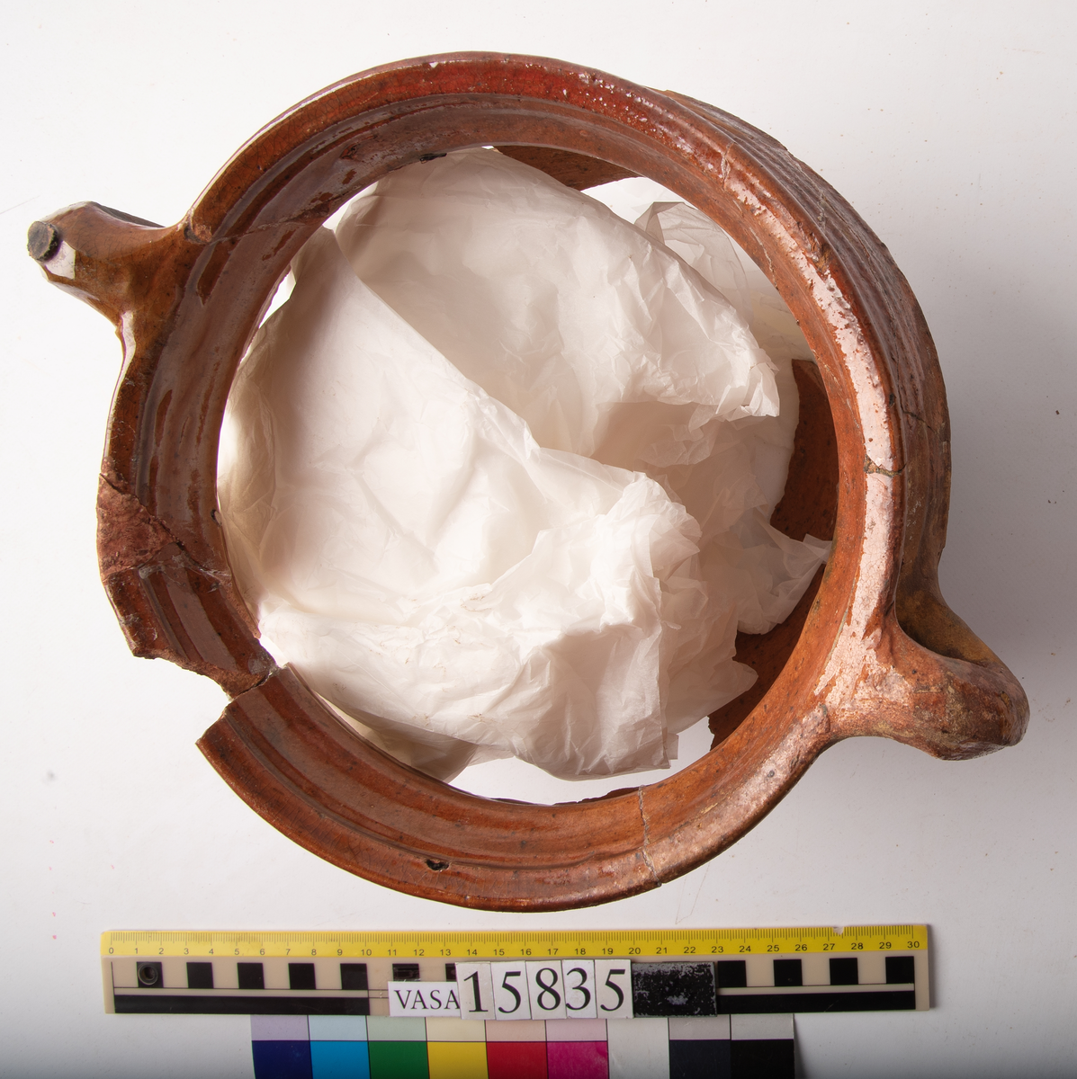 Kruka med två trindhänklar, ej komplett.
Krukan är fragmentarisk och sammansatt av ett flertal delar och bitar. Hela botten och delar av buken borta. Mynningsranden och bröstet nästan komplett. Mynningsvidd 190 mm. Förtjockad och profilerad mynningsrand med två trindhänklar som är ca 10 mm högre än mynningskanten.
På bröstet drejfåror. På skudervinkeln en ås och på bukkanten en ås.
Krukan är in- och utvändigt brunt blyglaserad, på buken mycket dåligt glaserad där den verkar sotig och sönderbränd.