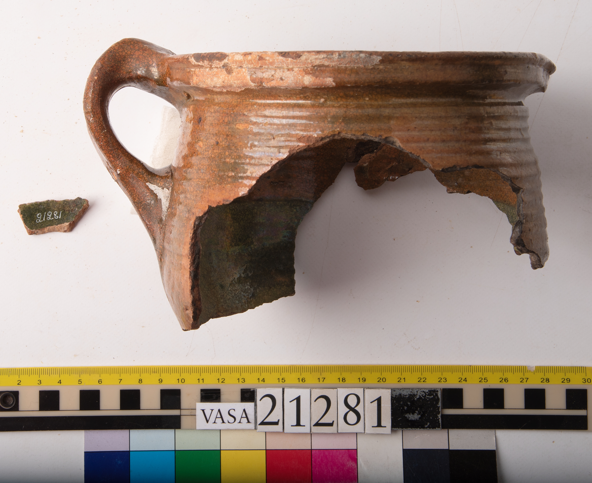 Kruka med bandhäkel och snås.
Krukan, fragmentariskt sammanhängande i en del med endast mynningsranden med bandkhänkeln och snåset samt skuldran intakt (komplett). Hela botten och större delen av buken borta.
Förtjockad och profilerad mynningsrand med snås och bandhänkel. Skuldran försedd med drejfåror ned till bukkanten. Krukan är på in- och utsidan blyglaserad brun med fläckvis svaga gröna skiftningar. Halva insidan nedåt har en klart markerad grön blyglasering.