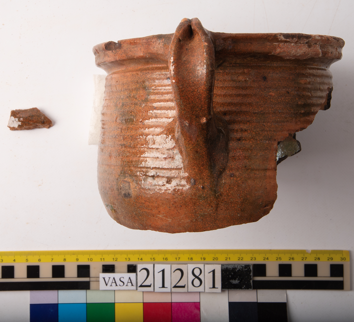 Kruka med bandhäkel och snås.
Krukan, fragmentariskt sammanhängande i en del med endast mynningsranden med bandkhänkeln och snåset samt skuldran intakt (komplett). Hela botten och större delen av buken borta.
Förtjockad och profilerad mynningsrand med snås och bandhänkel. Skuldran försedd med drejfåror ned till bukkanten. Krukan är på in- och utsidan blyglaserad brun med fläckvis svaga gröna skiftningar. Halva insidan nedåt har en klart markerad grön blyglasering.