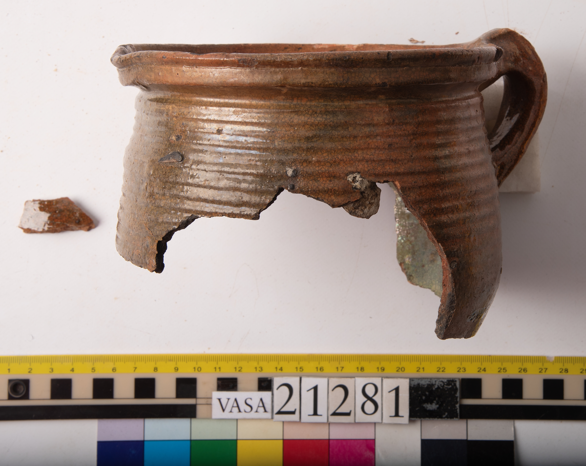 Kruka med bandhäkel och snås.
Krukan, fragmentariskt sammanhängande i en del med endast mynningsranden med bandkhänkeln och snåset samt skuldran intakt (komplett). Hela botten och större delen av buken borta.
Förtjockad och profilerad mynningsrand med snås och bandhänkel. Skuldran försedd med drejfåror ned till bukkanten. Krukan är på in- och utsidan blyglaserad brun med fläckvis svaga gröna skiftningar. Halva insidan nedåt har en klart markerad grön blyglasering.