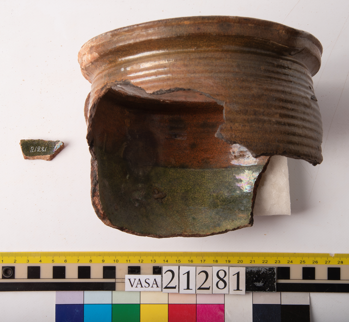 Kruka med bandhäkel och snås.
Krukan, fragmentariskt sammanhängande i en del med endast mynningsranden med bandkhänkeln och snåset samt skuldran intakt (komplett). Hela botten och större delen av buken borta.
Förtjockad och profilerad mynningsrand med snås och bandhänkel. Skuldran försedd med drejfåror ned till bukkanten. Krukan är på in- och utsidan blyglaserad brun med fläckvis svaga gröna skiftningar. Halva insidan nedåt har en klart markerad grön blyglasering.