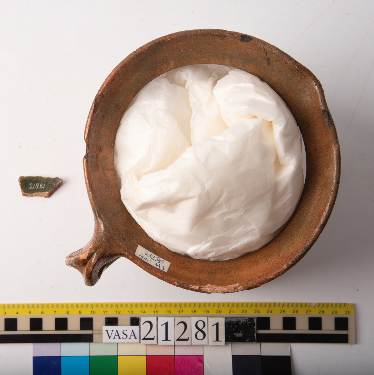 Kruka med bandhäkel och snås.
Krukan, fragmentariskt sammanhängande i en del med endast mynningsranden med bandkhänkeln och snåset samt skuldran intakt (komplett). Hela botten och större delen av buken borta.
Förtjockad och profilerad mynningsrand med snås och bandhänkel. Skuldran försedd med drejfåror ned till bukkanten. Krukan är på in- och utsidan blyglaserad brun med fläckvis svaga gröna skiftningar. Halva insidan nedåt har en klart markerad grön blyglasering.