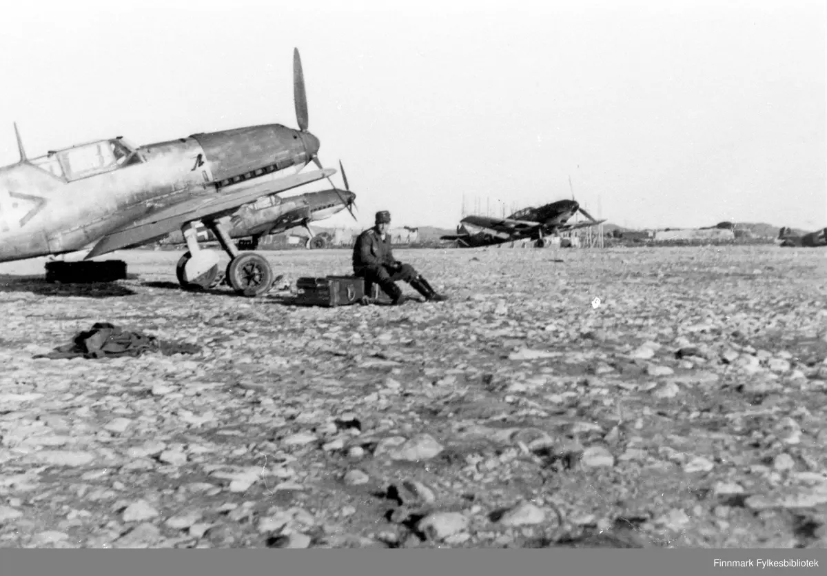 Messerschmitt Bf 109E-7 ved 2. gruppe av 5. jagerving (II./JG 5) parkert på plassen. Den nærmeste maskinen tilhører gruppens adjutant.

Informasjon fra publikum: "Bf 109E-7-flyet som står (foran) delvis skjult har merking <+, også det fra Gruppenstab II./JG 5. Også dette viser en mørkere motordeksel, mens den nedre delen under eksosutslippene trolig fortsatt er gul på begge disse flyene. Dette som identifikasjon for egne styrker og luftvern."