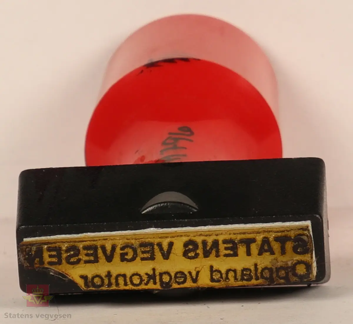 Stempel av plast og stempelplaten er av gummi. Rektangulært stempel med rødt håndtak som er avlangt og som utvider seg. Det er en preget logo på toppen av gjenstand av ukjent navn og opprinnelse.

