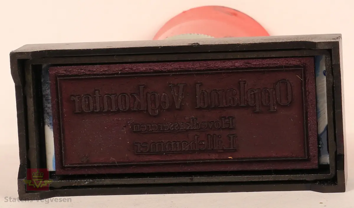 Stempel av plast og stempelplaten er av gummi. Rektangulært, svart stempel med rødt håndtak. På framsiden av stempel står stempel typen i gul skrift. Stempelplaten er blåfarget pga. blekk. 