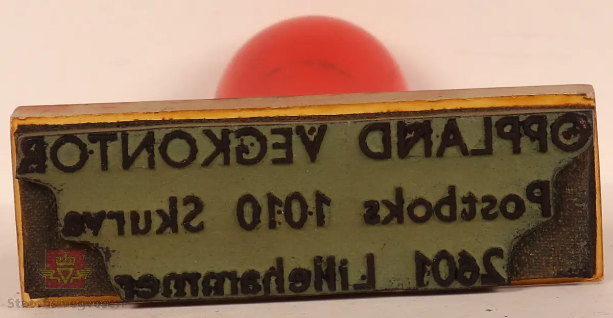 Stempel av tre, plast og gummi. Rektangulært formet med avlangt håndtak. Håndtaket er rødt og stempelplaten gjennomsiktig øverst, med grå stempelplate under og svart tekst. 