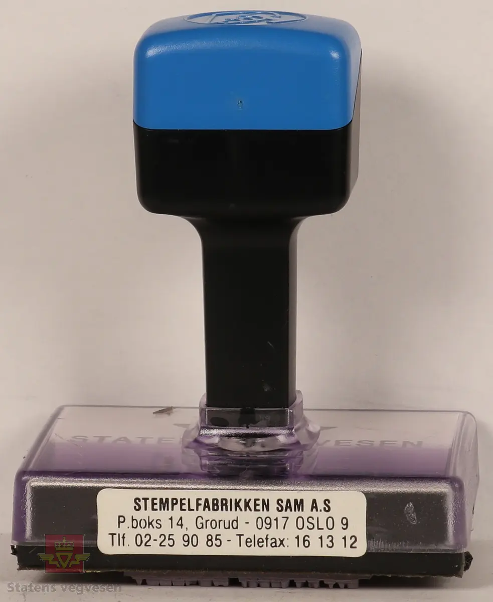 Blått, hvitt og svart stempel i plast. Firkantet håndtak med stempellogo på toppen. Stempelplaten er gjennomsiktig øverst med hvit bakgrunn hvor det står tekst om hvordan stempelet skal se ut. Stempelplaten under er grå med vegvesenets emblem og tekst. 