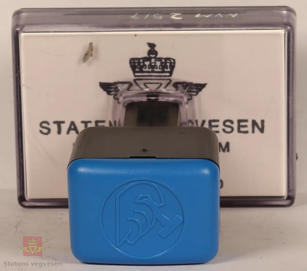 Blått, hvitt og svart stempel i plast. Firkantet håndtak med stempellogo på toppen. Stempelplaten er gjennomsiktig øverst med hvit bakgrunn hvor det står tekst om hvordan stempelet skal se ut. Stempelplaten under er grå med vegvesenets emblem og tekst. 
