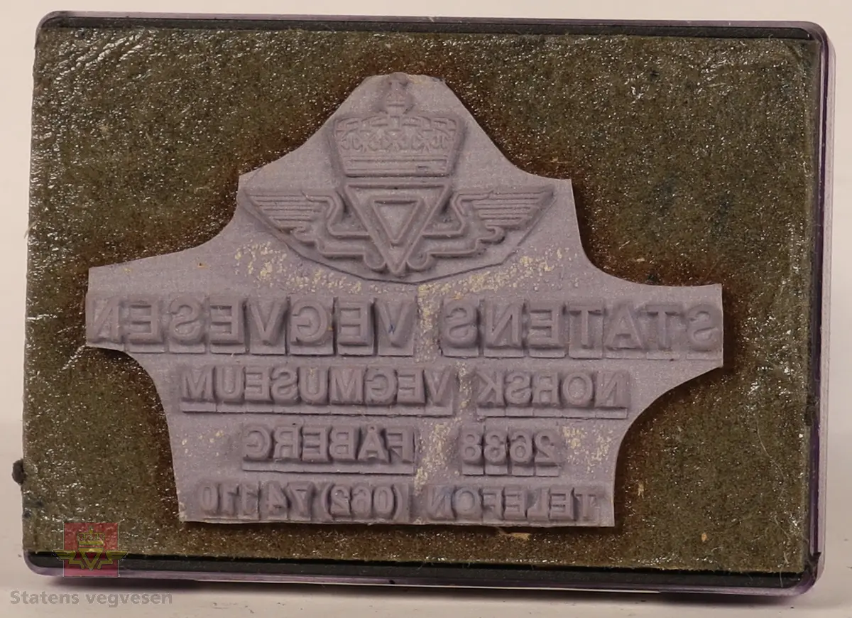 Blått, hvitt og svart stempel i plast. Firkantet håndtak med stempellogo på toppen. Stempelplaten er gjennomsiktig øverst med hvit bakgrunn hvor det står tekst om hvordan stempelet skal se ut. Stempelplaten under er grå med vegvesenets emblem og tekst. 