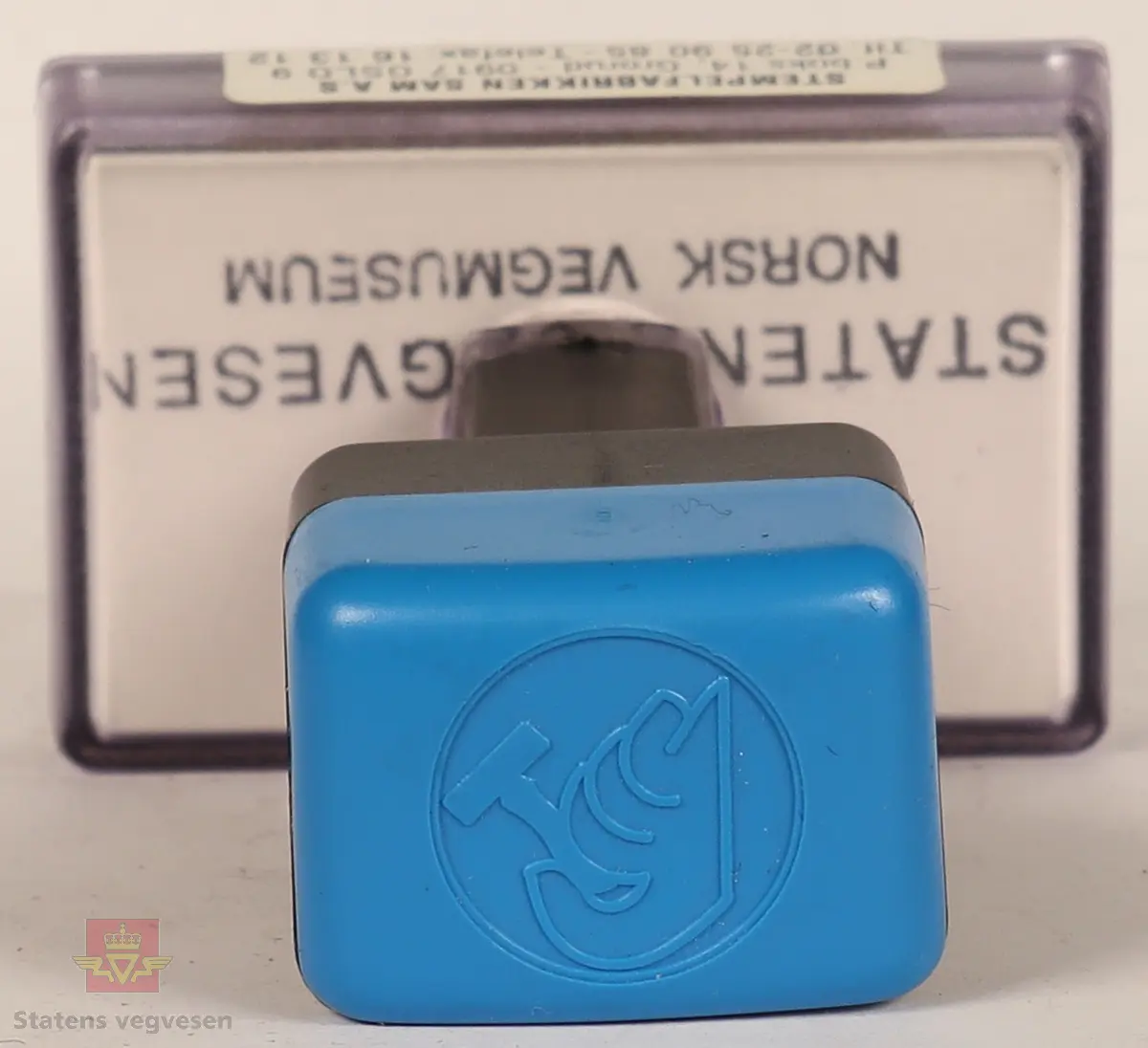 Blått, hvitt og svart stempel i plast. Firkantet håndtak med stempellogo på toppen. Stempelplaten er gjennomsiktig øverst med hvit bakgrunn hvor det står tekst om hvordan stempelet skal se ut. Stempelplaten under er grå med vegvesenets emblem og tekst. 
