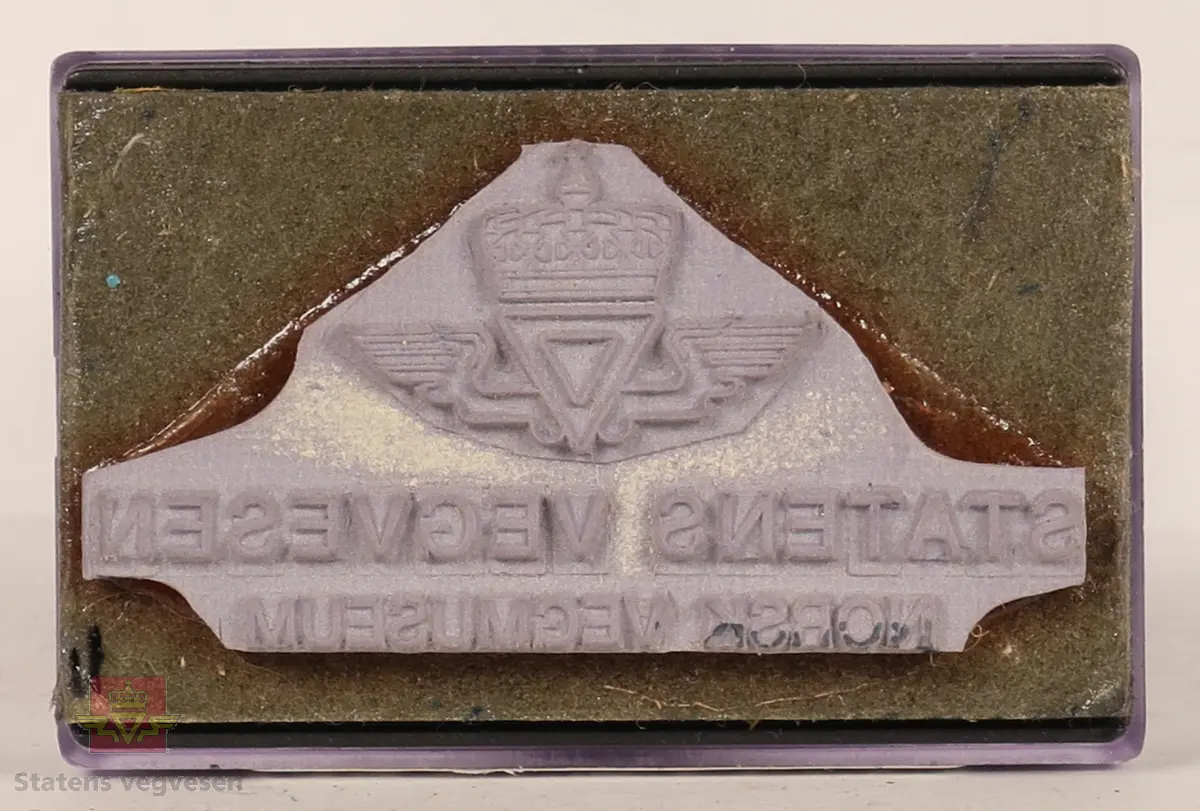 Blått, hvitt og svart stempel i plast. Firkantet håndtak med stempellogo på toppen. Stempelplaten er gjennomsiktig øverst med hvit bakgrunn hvor det står tekst om hvordan stempelet skal se ut. Stempelplaten under er grå med vegvesenets emblem og tekst. 