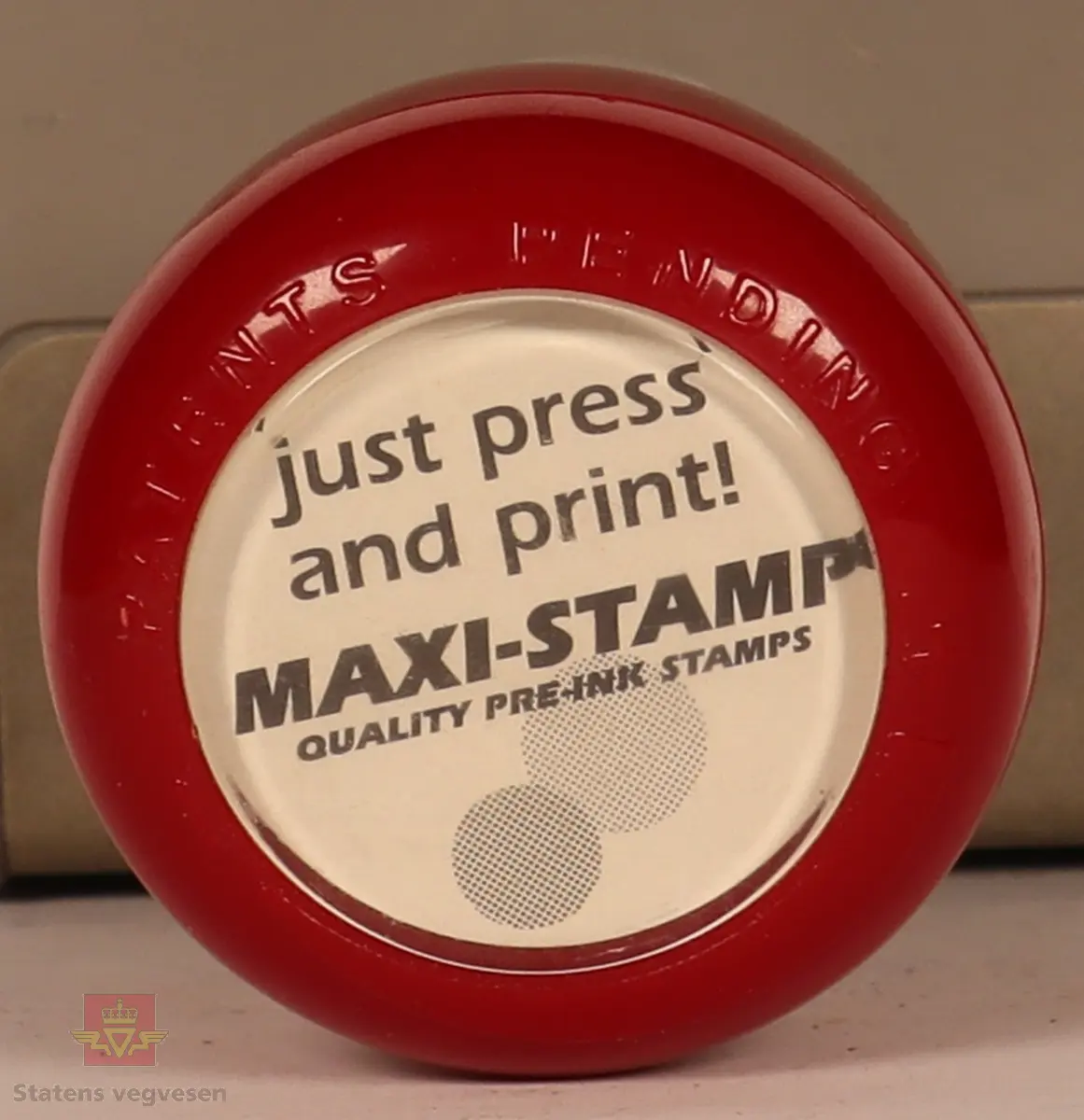 Rødt, grått, hvitt og svart stempel i plast. Firkantet med rundt håndtak. Stempel puten er tørket og har vegvesenets logo med tekst. 