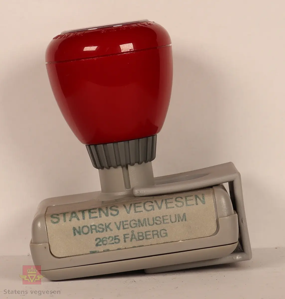 Rødt, grått, hvitt og svart stempel i plast. Firkantet med rundt håndtak. Stempel puten er tørket og har vegvesenets logo med tekst. 