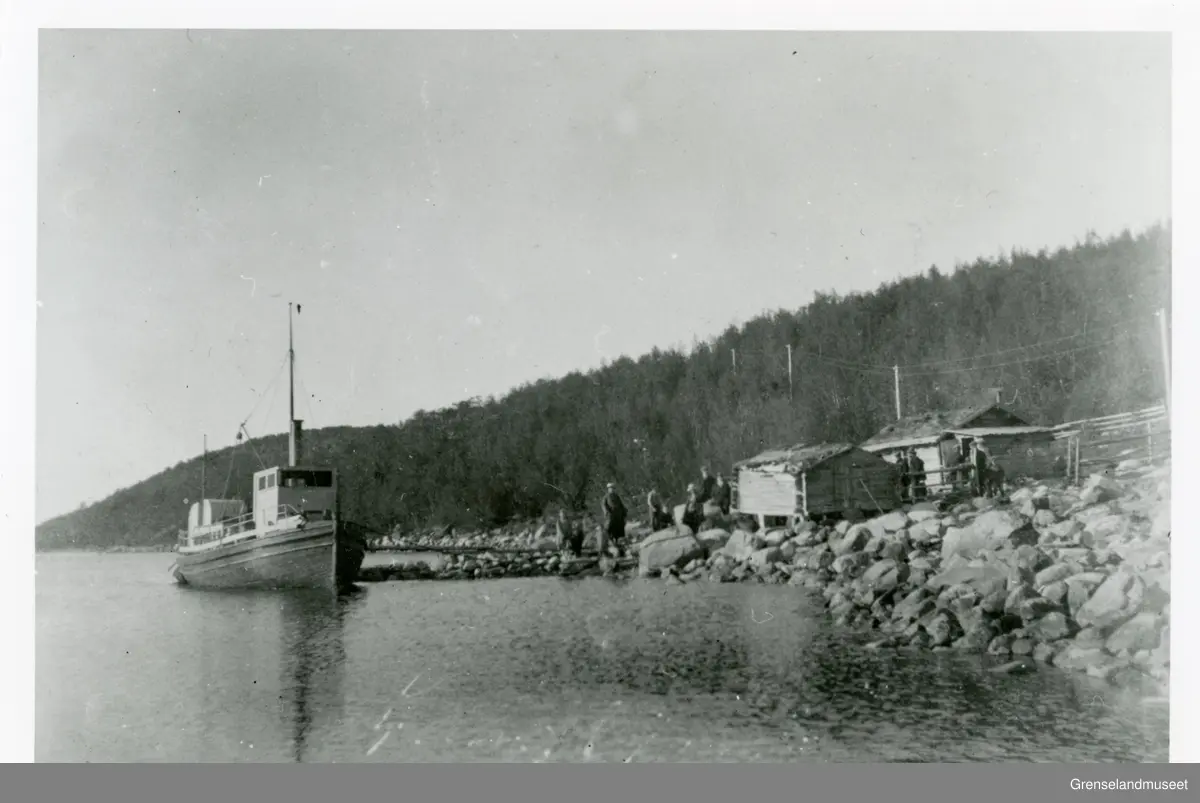 Motorbåten "Langfjorden" fortøyd til kaia. En gruppe mennesker står ved foten av kaia, nært noen små bygninger. 