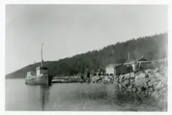 Motorbåten "Langfjorden" fortøyd til kaia. En gruppe mennesker står ved foten av kaia, nært noen små bygninger.