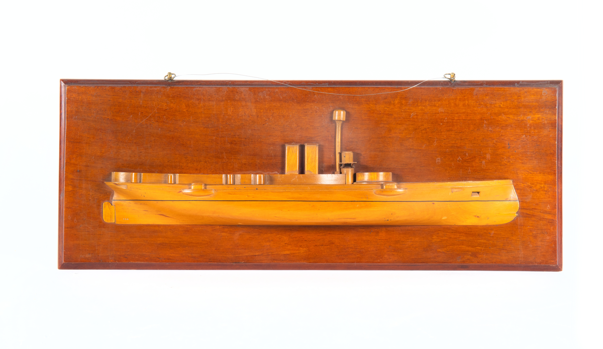 Fartygsmodell med roder, mast, 2 fyrkantiga skorstenar och  brygga. Fästad til väggplan.